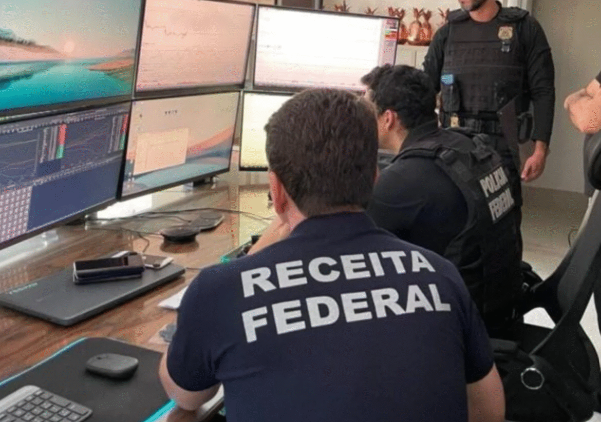 Fotografia em close-up de um Agente da Receita Federal virado de costas com a camiseta da RFB, concentrado em cálculos em uma mesa com papéis e uma calculadora, estudando a legislação sobre salários e impostos.